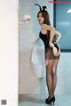 A woman in a black bodysuit and stockings standing next to a urinal.